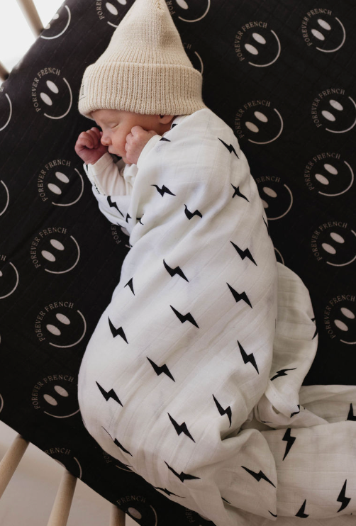 A newborn baby lies peacefully in a crib, swaddled in the reversible Muslin Quilt | ff Smile Black & White Bolt from forever french baby, with black lightning bolt patterns made from 100% muslin cotton. The baby is wearing a knitted beige hat, and the crib mattress features a black cover with white smiley faces and the text "FOREVER FRENCH BABY.