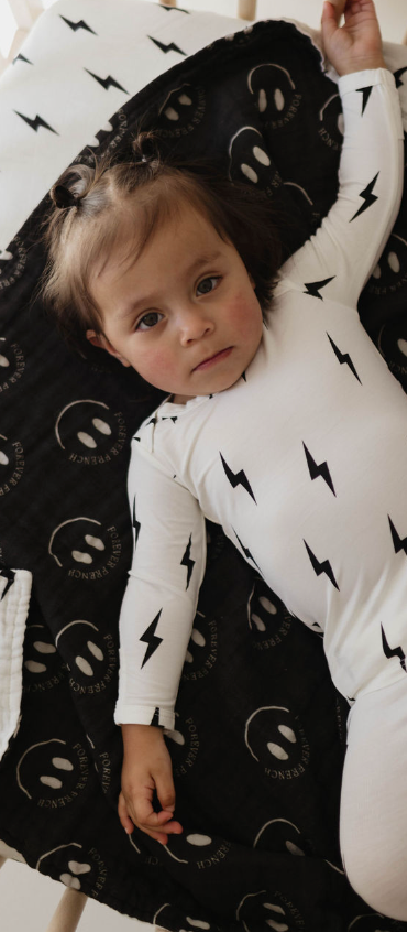 A baby dressed in a white onesie with black lightning bolt patterns lies on a forever french baby Muslin Quilt, featuring the ff Smile Black & White Bolt design. The quilt is made from 100% Muslin Cotton and adorned with white circular designs and illustrations of smiling faces. The baby looks up with a calm expression, surrounded by a cozy and monochromatic setting.