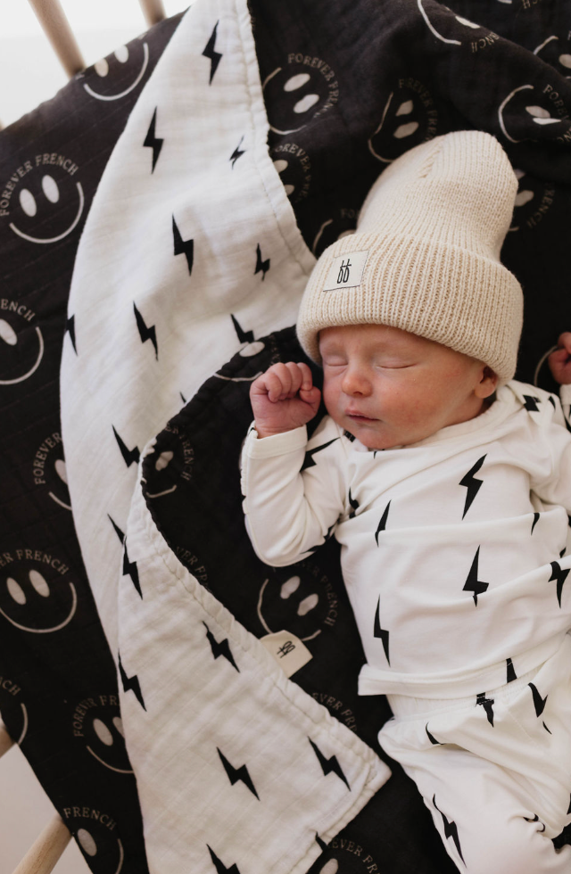 A sleeping baby is dressed in a white outfit with black lightning bolt patterns and a beige knit hat. The baby is nestled under the Muslin Quilt | ff Smile Black & White Bolt from forever french baby, featuring smiley faces and lightning bolts. The environment looks cozy and peaceful.