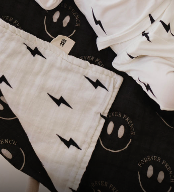 A close-up photo showcases 100% muslin cotton fabric with white designs on a black background, featuring smiley faces and the text "FOREVER FRENCH" alongside lightning bolts. A reversible Muslin Quilt from forever french baby, named "ff Smile Black & White Bolt," adorned with black lightning bolts on white cloth, partially overlaps the black fabric.