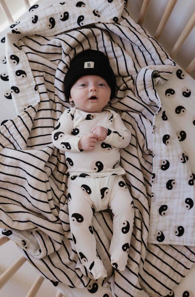 A baby wearing a black beanie and a white outfit with yin-yang symbols lies on a patterned blanket in a crib. The reversible muslin quilt and crib sheet, from forever french baby, feature matching Black & White Yin Yang and striped designs. Made from 100% muslin cotton, the Muslin Quilt | Black & White Yin Yang / Stripe ensures comfort as the baby rests with hands together, eyes open.
