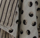 Close-up of two pieces of fabric. The left fabric, part of the "Muslin Quilt | Black & White Yin Yang / Stripe" by forever french baby, is crafted from 100% muslin cotton and features a beige base with black vertical stripes. The right fabric showcases a pattern of black and white yin-yang symbols. Both fabrics appear to have soft, woven textures.