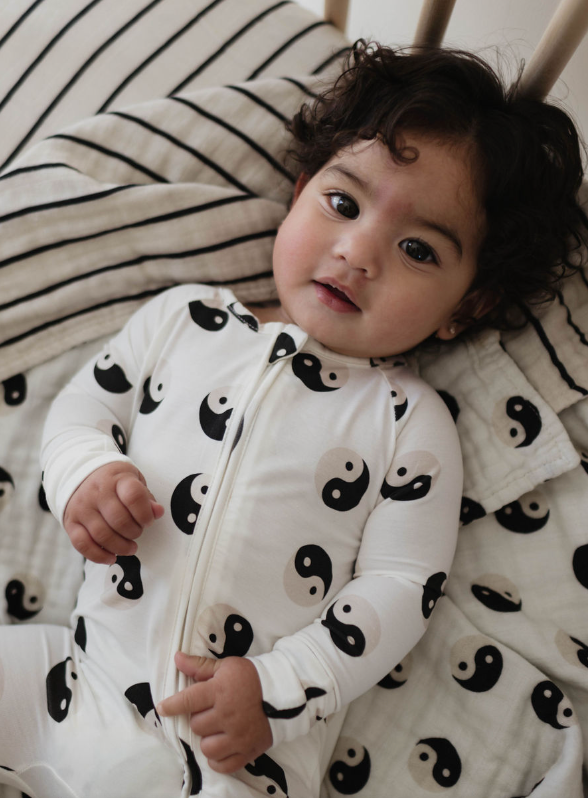 A baby with curly hair is lying on the Muslin Quilt | Black & White Yin Yang / Stripe by forever french baby, made from 100% muslin cotton, and wearing a white onesie adorned with black and white yin-yang symbols. The baby gazes directly at the camera with a relaxed expression.