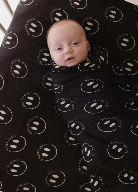A baby is swaddled in a super-soft muslin blanket adorned with white smiley faces and the words "FOREVER FRENCH." The baby is lying on the Muslin Crib Sheet | Charcoal & White ff Smile by forever french baby, looking up with wide eyes.