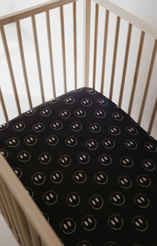 A wooden crib with vertical slats contains a mattress covered with the Muslin Crib Sheet | Charcoal & White ff Smile from forever french baby, adorned with smiling faces and the words "Happy Friends" in white text. Crafted from super-soft muslin, this design brings a cheerful and playful touch to the nursery setting.