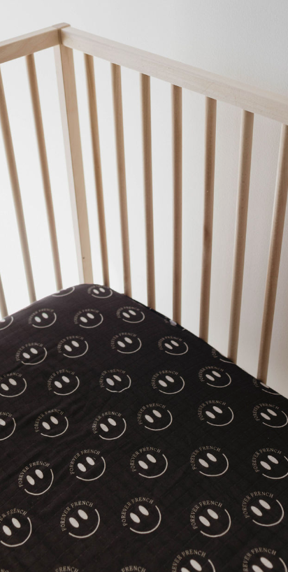 A wooden baby crib with vertical slats features the Muslin Crib Sheet | Charcoal & White ff Smile from forever french baby. The super-soft muslin sheet is adorned with white smiley faces and the text "HELLO FRENCH" in a repeating pattern. The minimalist setting emphasizes the crib and its charming bedding.