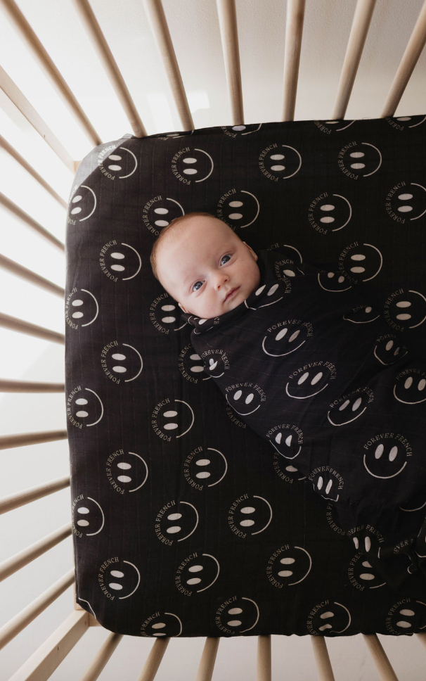 A baby lies in a crib, swaddled in a black blanket adorned with white smiley faces and the text "FOREVER FRIENDS." The crib mattress features a Muslin Crib Sheet | Charcoal & White ff Smile from forever french baby, showcasing the same cheerful pattern. Surrounded by super-soft muslin comfort, the baby looks at the camera with wide eyes.