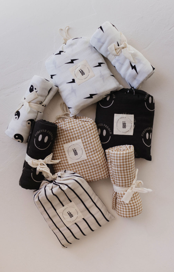 Forever French Baby's Muslin Swaddle collection, featuring the Black & White Lightning Bolt design among other charming patterns like smiley faces, gingham check, stripes, and yin-yang symbols, provides comforting softness for your baby. Each swaddle is neatly rolled and folded, tied with a fabric bow tie for a delightful touch.