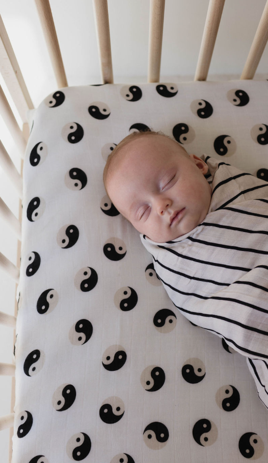 A baby is peacefully sleeping in a crib, swaddled in a blanket with black and white stripes. The Muslin Crib Sheet | Black & Ivory Yin Yang by forever french baby decorates the mattress, adding to the serene scene with its intricate pattern.