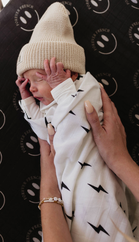A newborn baby, wrapped in a "Muslin Swaddle | Black & White Lightning Bolt" by forever french baby, wears a cream knit hat. The baby is being held gently by two adult hands. The background features a black cloth with white circular logos and the text "FOREVER FRENCH.