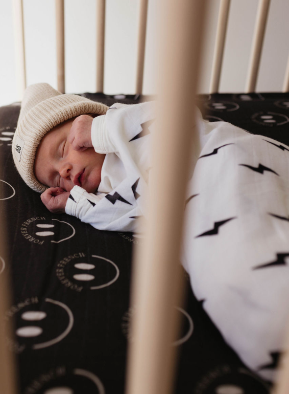 A newborn baby sleeps peacefully in a crib, wearing a tan knit hat and a white onesie with a black lightning bolt pattern. Wrapped in the forever french baby Muslin Swaddle | Black & White Lightning Bolt for added comfort, the crib mattress has a black cover decorated with white smiley faces and text. Soft light filters into the cozy scene.