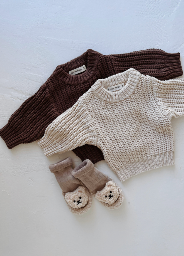 Two children’s cotton sweaters from forever french baby, including the Cream | Knit Sweater, are laid flat on a white surface. Below them is a pair of beige baby socks with plush animal faces on the toes.