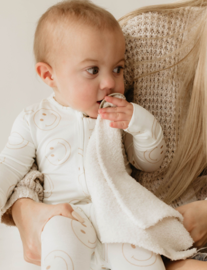 A baby dressed in a white onesie with smiling face prints is sitting on a woman's lap, chewing on the corner of the softest "Lovey | Golden Grid" from forever french baby. The woman, whose face is not visible, is wearing a knitted beige sweater made from microfiber feather yarn, supporting the baby gently.