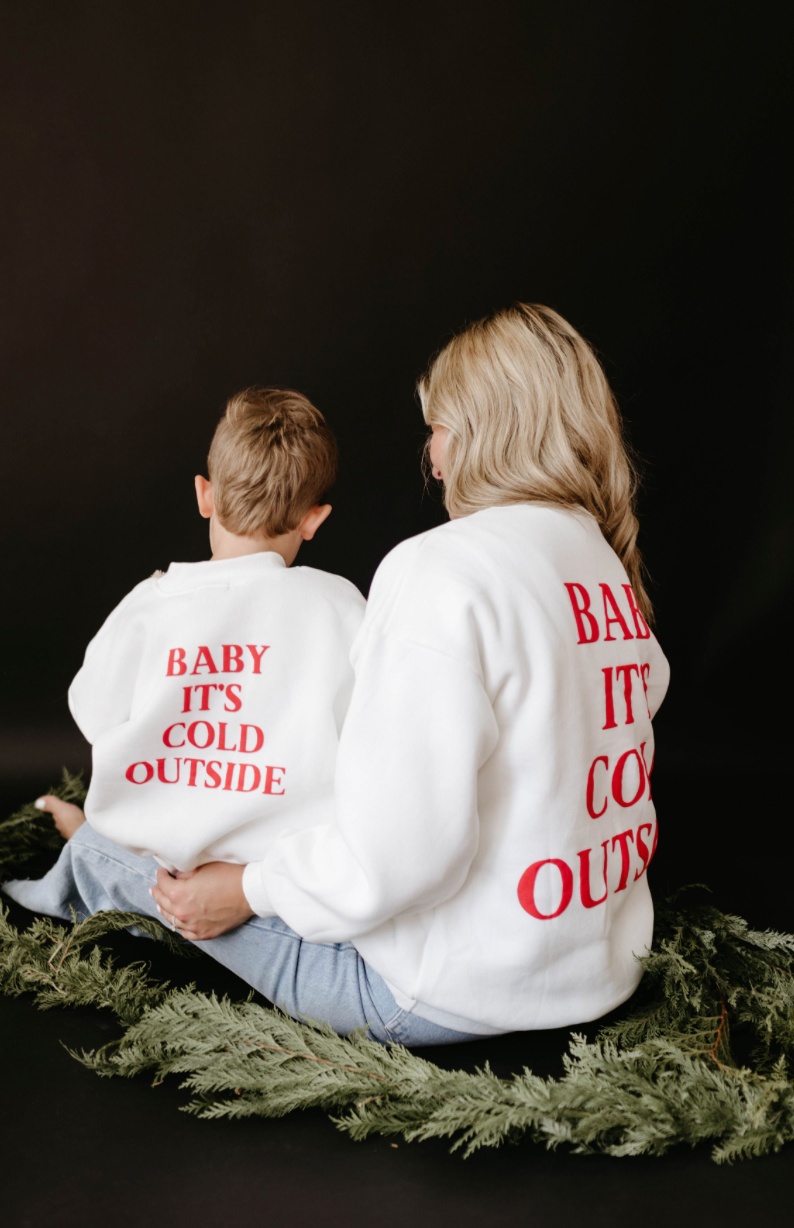 A woman and child sit on evergreen branches against a dark background, both wearing oversized white crewnecks featuring the red text "Baby It's Cold Outside" on the back, perfectly capturing the spirit of holiday apparel and ideal for creating family matching outfits. The child's sweatshirt is from forever french baby's 'Child Sweatshirt | Baby It's Cold Outside' collection.