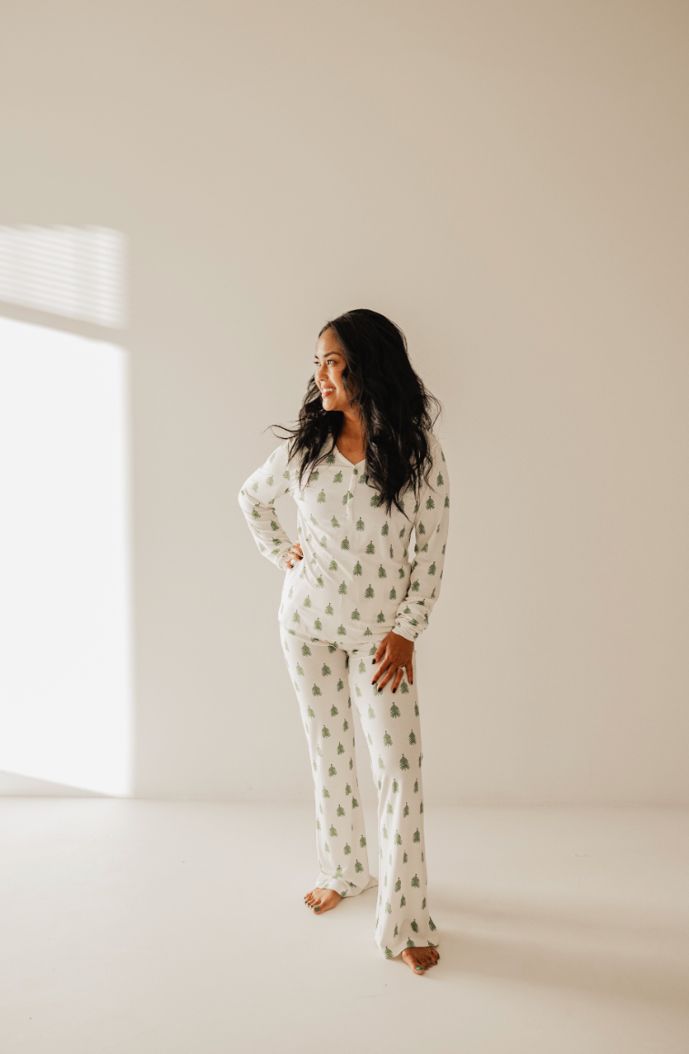 In a sunlit room with plain beige walls, a person with long dark hair stands barefoot wearing the cozy "O Christmas Tree" bamboo pajamas by forever french baby, featuring rollover cuffs and a small green tree pattern, as they gaze to the left with a relaxed expression.