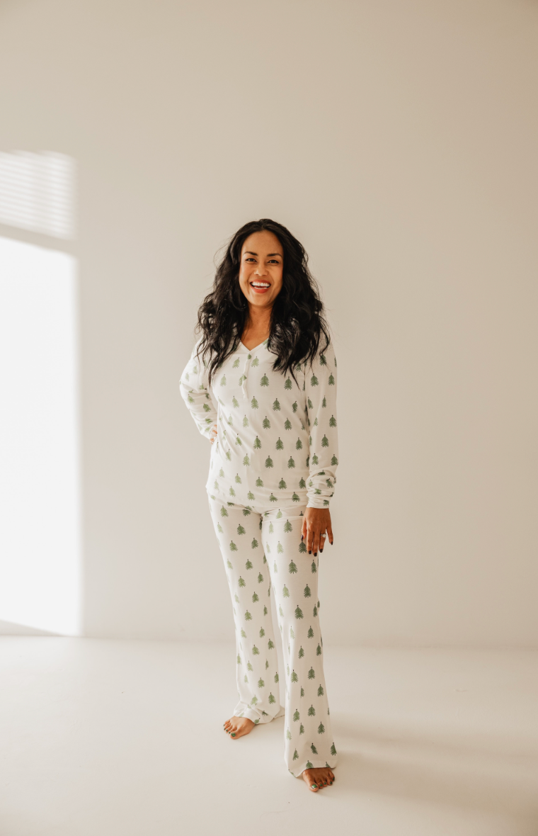 A person with long hair is smiling on a bright, sunlit floor, embracing the morning in cozy O Christmas Tree pajamas from forever french baby. The plain white background is softly illuminated by window light.