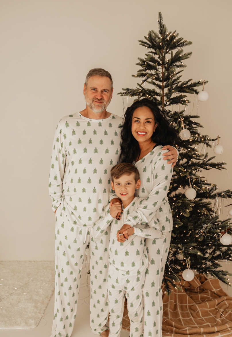 A family of three, dressed in forever french baby O Christmas Tree bamboo pajamas with rollover cuffs, poses warmly by a decorated tree with white ornaments and lights. They stand on a beige rug against a plain background, their smiles illuminating the holiday spirit.