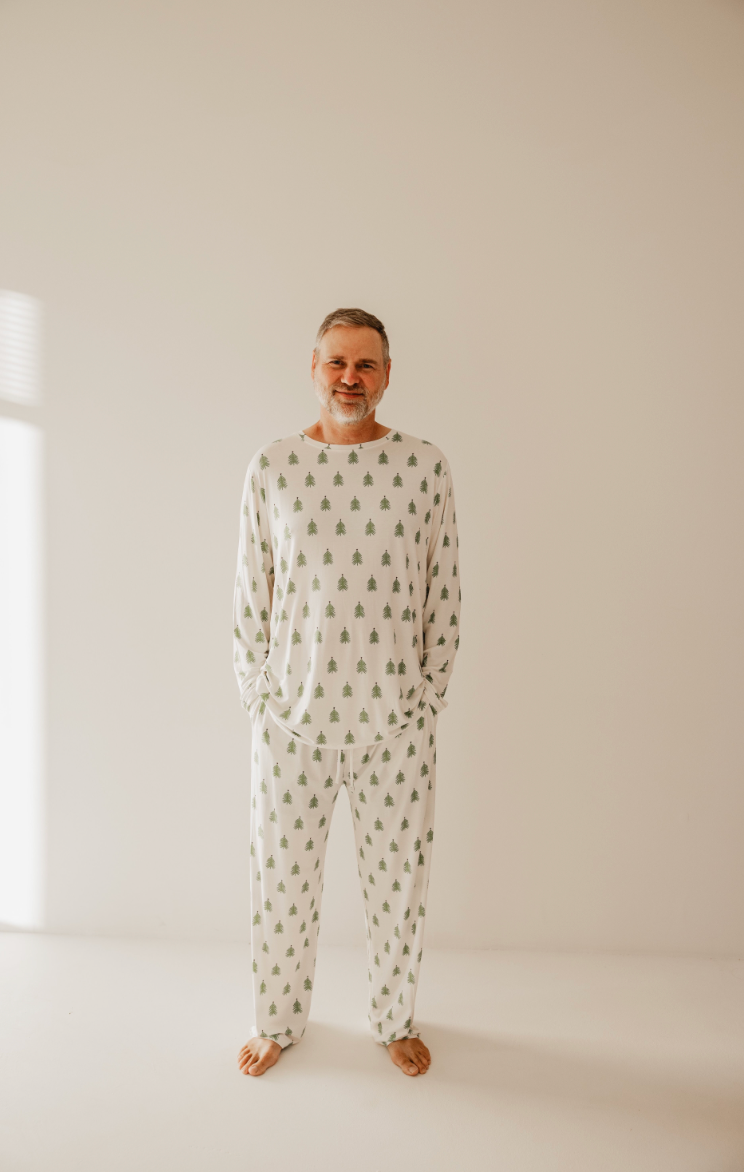 A man stands barefoot in a minimal room, wearing cozy "O Christmas Tree" bamboo pajamas by forever french baby, featuring small green tree patterns, softly lit from the left.