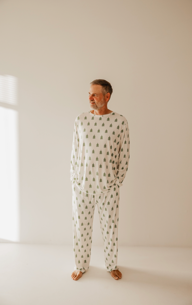 A man stands against a plain background wearing forever french baby's O Christmas Tree Men Bamboo Pajamas adorned with small trees. Soft window light casts gentle shadows, and with his hands in his pockets, he exudes relaxation and cozy comfort.