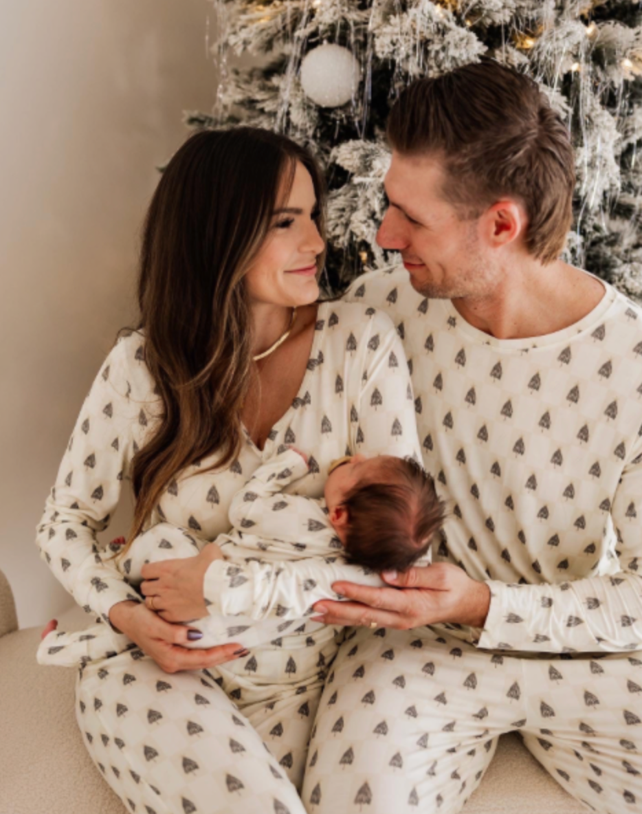 A couple in matching Vintage Christmas Deck the Halls Checkerboard pajamas from forever french baby holds a Forever French Baby near a decorated tree. They sit closely, sharing warm gazes. The scene with its soft fabrics and gentle smiles exudes a cozy, festive atmosphere.