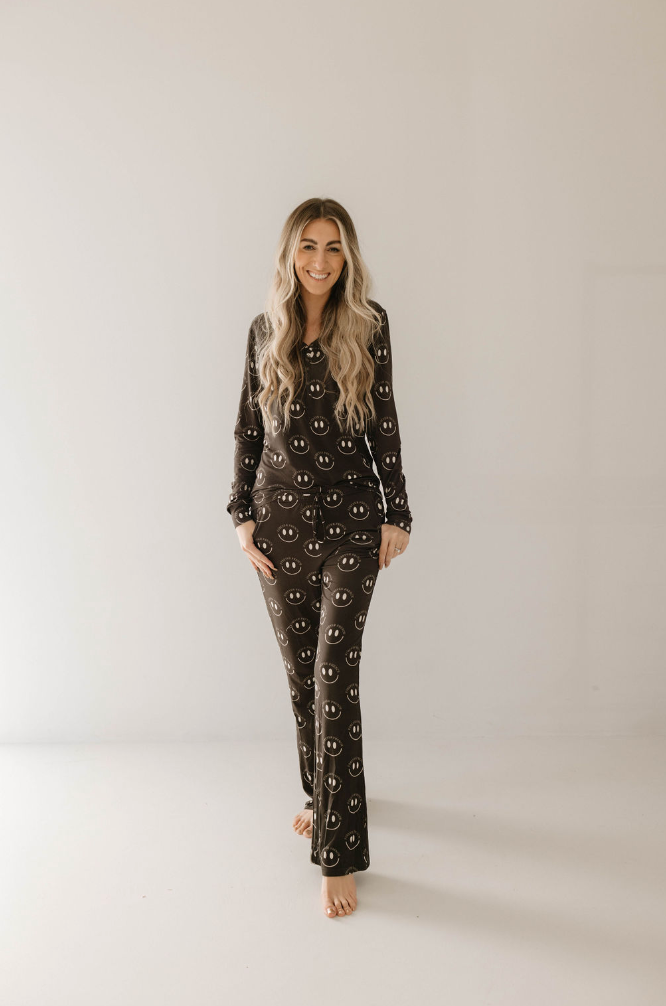 A person with long, wavy hair stands barefoot against a plain background, smiling. They are wearing the Women's Bamboo Pajamas in Charcoal & White ff Smile by forever french baby. Made from hypo-allergenic bamboo, these pajamas offer year-round comfort and feature a matching dark set adorned with a pattern of smiley faces. Their hands are in their pockets, and they exude a relaxed, happy demeanor.