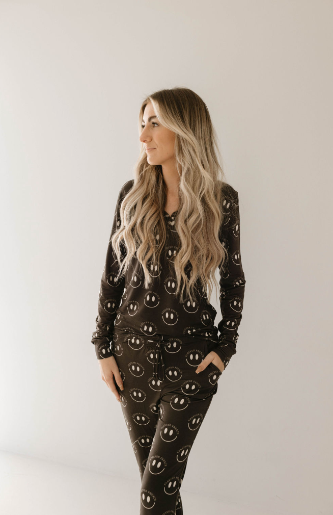 A person with long, wavy hair stands against a plain white background, wearing a dark matching set of hypo-allergenic Women's Bamboo Pajamas, Charcoal & White ff Smile by forever french baby. The individual looks to the side with their hands in their pockets, enjoying year-round comfort.