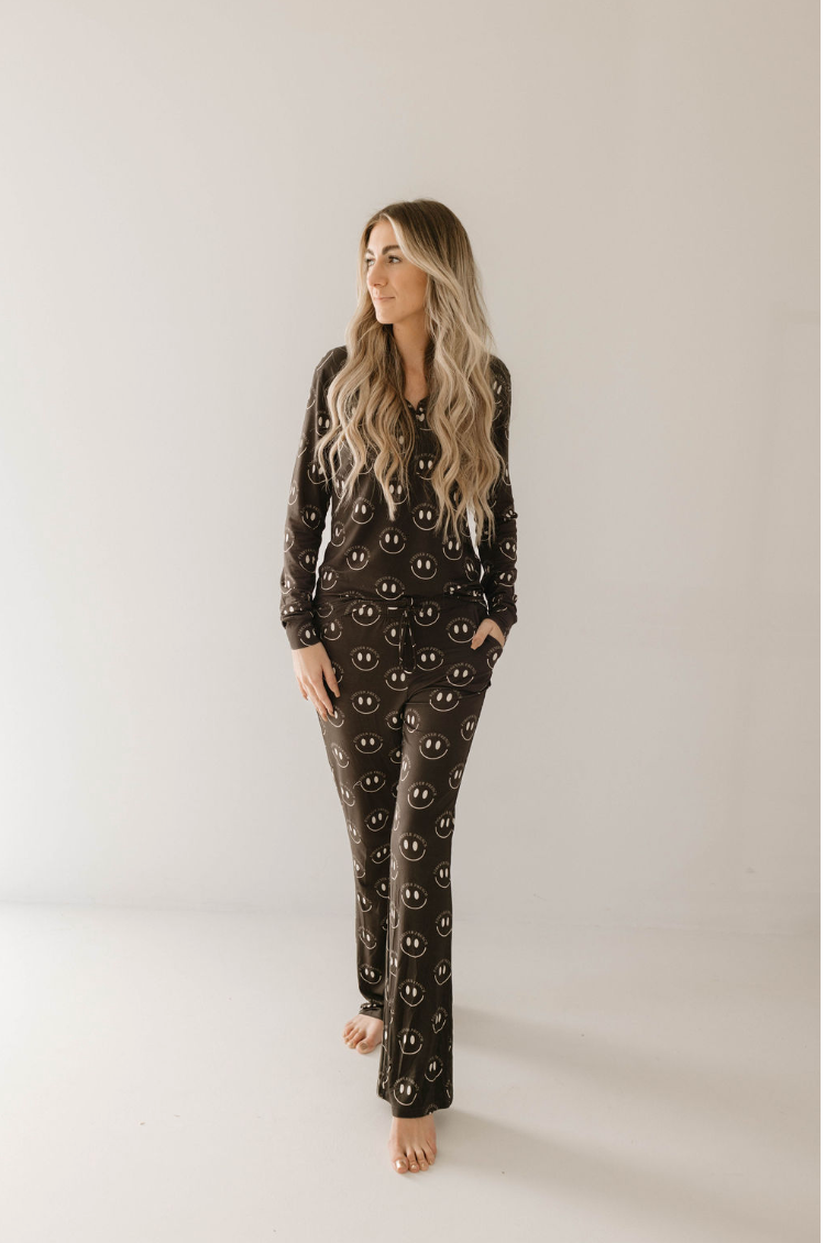A person with long, wavy hair stands barefoot against a plain background, wearing the hypo-allergenic Women's Bamboo Pajamas by forever french baby, featuring a charcoal and white smiley face pattern. The dark-colored set offers year-round comfort as the person has one hand in their pocket and gazes to the side.
