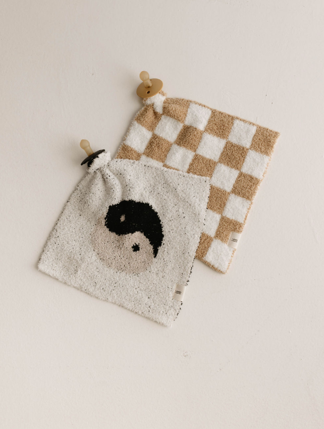 Two soft cloths with pacifiers attached are laid on a light-colored surface. One cloth, named the Lovey | Original Checkerboard from forever french baby, boasts a beige and white checkered pattern made from microfiber feather yarn. The other features an elegant yin-yang symbol in black and beige—perfect little loveys from Forever French Baby.