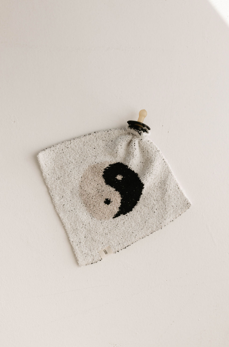 The Lovey | Yin-Yang Black & White from forever french baby is a square-shaped knitted cloth made from microfiber feather yarn, featuring a black and white yin-yang symbol at its center and a pale-colored pacifier attached to one corner. This softest lovey rests on a plain, light-colored surface.