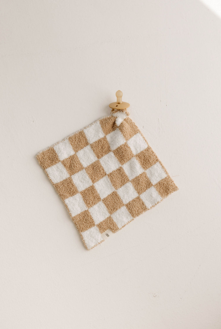 The Lovey | Original Checkerboard from forever french baby, featuring a beige and white checkered microfiber feather yarn cloth, is displayed on a minimalistic wooden round wall hook against a plain white wall. The textured fabric is neatly draped over the hook, adding a simple, natural touch to the overall scene. Perfect for any Forever French Baby nursery.