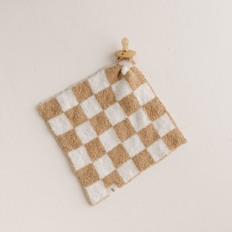 The Lovey | Original Checkerboard from forever french baby, featuring a beige and white checkered microfiber feather yarn cloth, is displayed on a minimalistic wooden round wall hook against a plain white wall. The textured fabric is neatly draped over the hook, adding a simple, natural touch to the overall scene. Perfect for any Forever French Baby nursery.