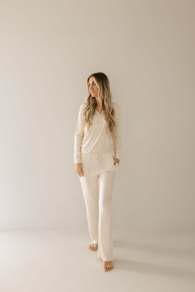 A woman with long, wavy hair is wearing the Golden Grid Women's Bamboo Pajamas by forever french baby. She is standing barefoot in a neutral-toned room, looking slightly to her right with a relaxed expression. Her hands are casually in her pockets, embodying the cozy elegance of Forever French Baby.