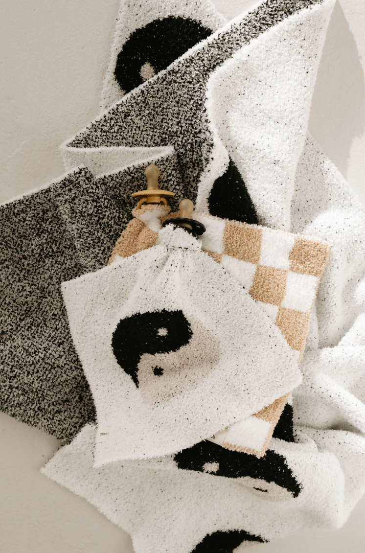 A collection of textured, patterned towels from forever french baby hangs against a wall. Among them, the Lovey | Original Checkerboard towel stands out with its black and white speckles, checkered designs, and abstract face prints. Wooden towel hooks are partially visible at the top, adding to the artistic and decorative arrangement.