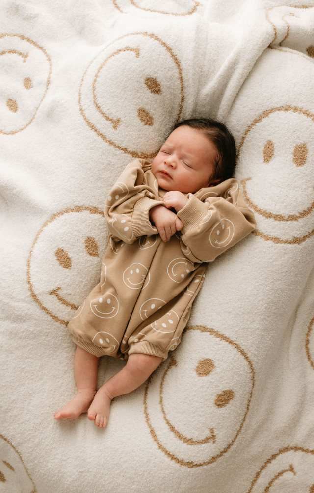 A newborn sleeps peacefully on a white blanket with tan smiley faces, wearing the forever french baby "Just Smile" long sleeve romper featuring white smiley faces. The baby's arms are crossed over their chest.