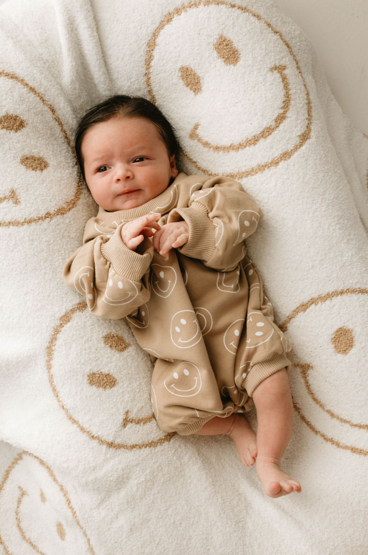 A dark-haired baby lies on a soft white blanket with tan smiley faces, wearing the forever french baby Just Smile long sleeve romper decorated with white smiley face patterns, and gazes upward gently.