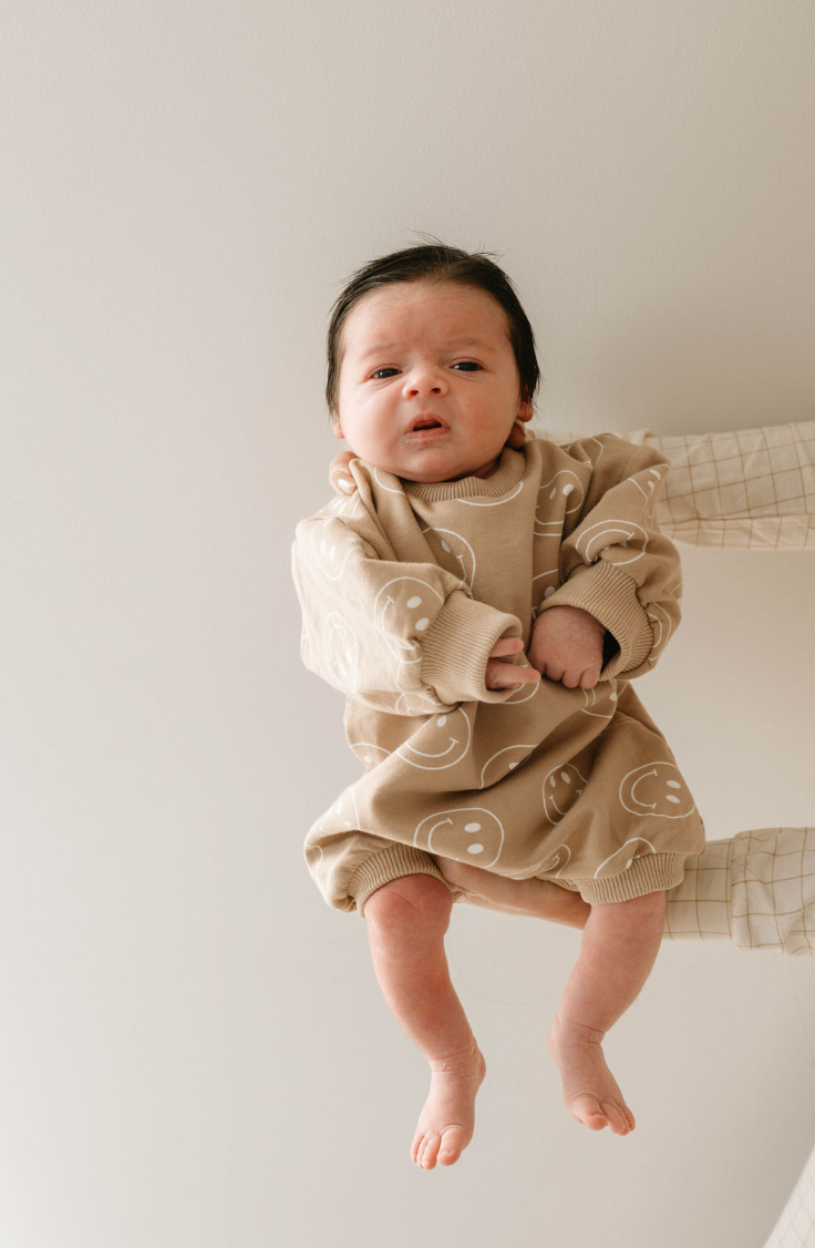 A baby with dark hair and a neutral expression is held upright, wearing the forever french baby Just Smile | Long Sleeve Romper—beige, organic cotton, with white smiley faces—against a plain light background.