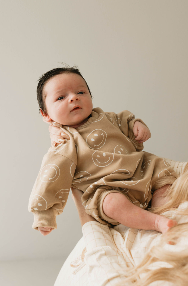 A baby with dark hair wears the beige "Just Smile" long sleeve romper by forever french baby, patterned with white smiley faces, while being held by an adult against a light, plain background.