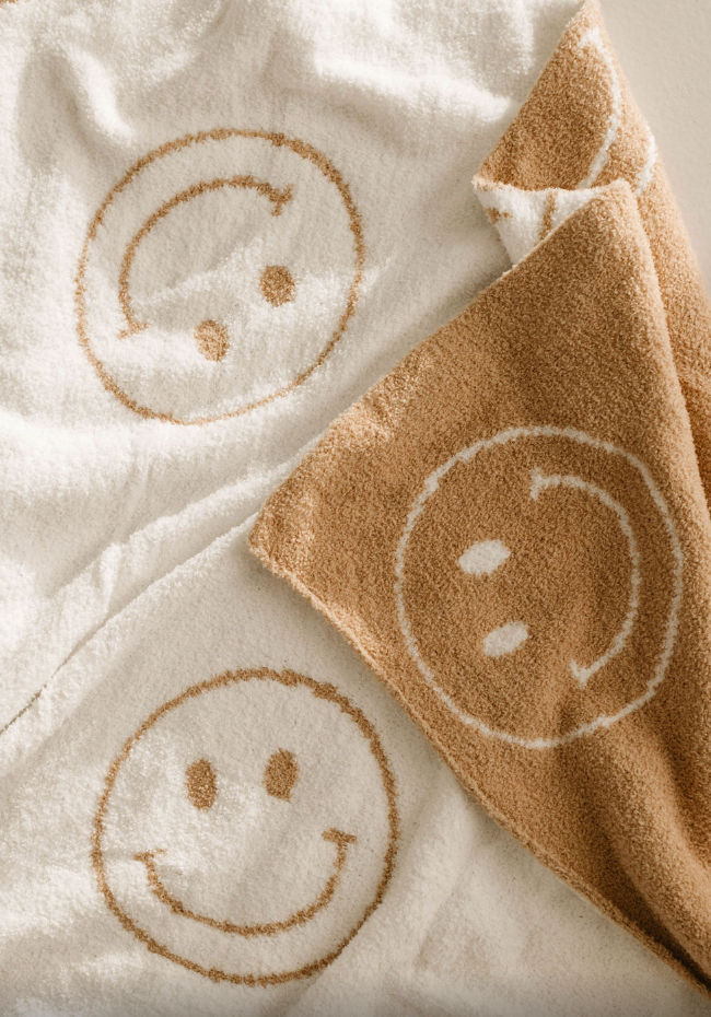 A close-up of two Plush Blankets by forever french baby, from the Original Just Smile line, in white and tan soft microfiber feather yarn with large contrasting smiley face designs, slightly overlapping and softly textured.