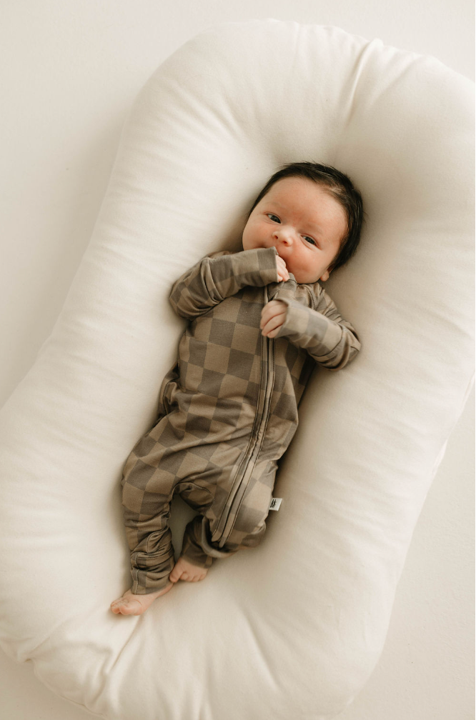 A baby with dark hair lies on a white, cushioned surface. The baby is wearing the "Bamboo Zip Pajamas | Faded Brown Checkerboard" from forever french baby, and one hand is near the mouth while the baby gazes upwards.
