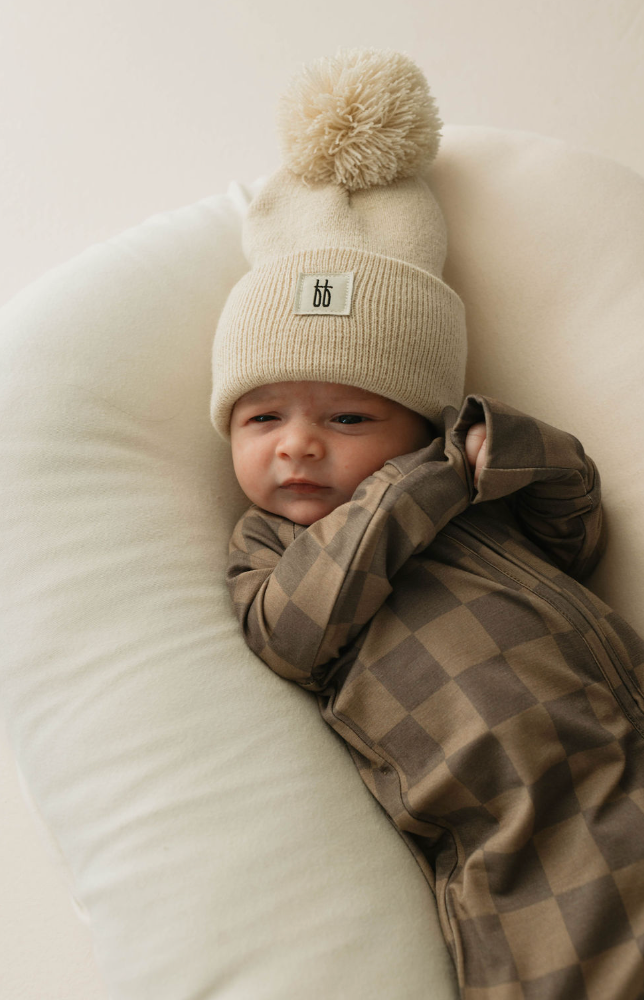 A baby is lying on a soft, white cushion while wearing a breathable beige knit hat with a large pom-pom and the Bamboo Zip Pajamas in Faded Brown Checkerboard by "forever french baby." The baby's hands are near their face, and they look calm and slightly curious.