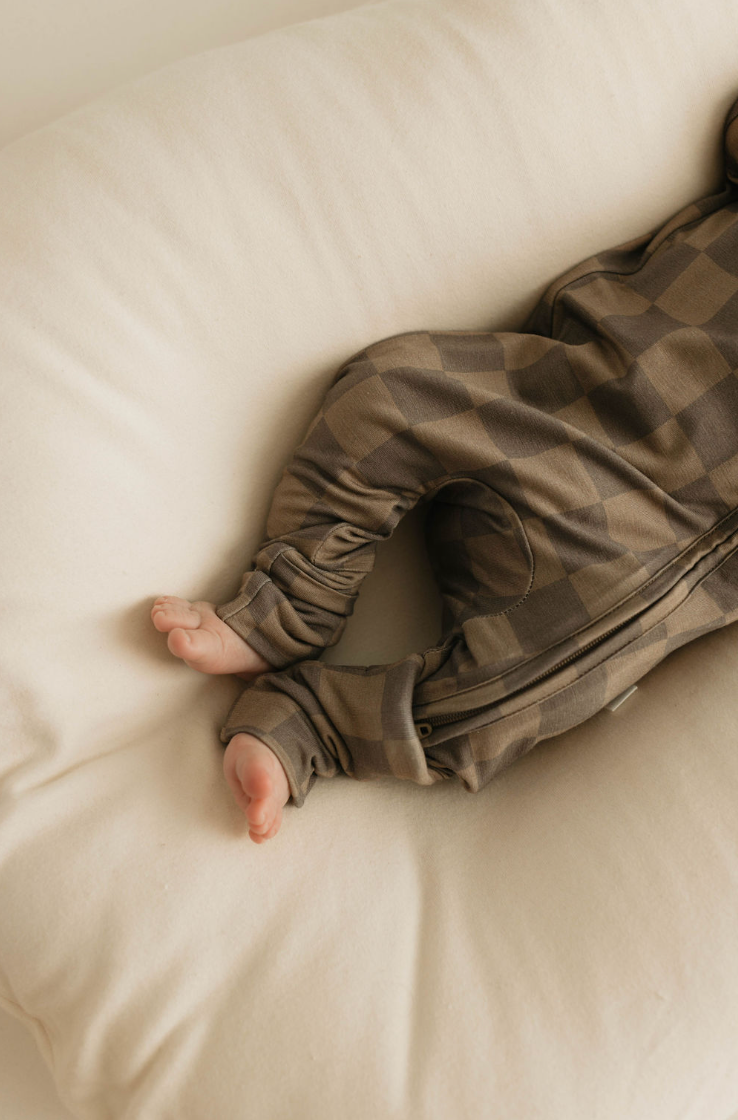 Close-up of a baby's legs and feet resting on a light-colored cushion. The baby is dressed in the Bamboo Zip Pajamas in Faded Brown Checkerboard, made from hypo-allergenic bamboo clothing by forever french baby. The baby's feet are slightly crossed over each other.