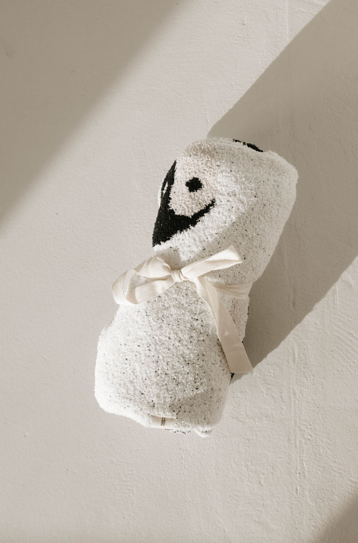 A Plush Blanket | Black & White Yin Yang from forever french baby, textured and velvety-rich, is rolled and tied with a beige ribbon on a light surface. Made from microfiber feather yarn, it features a black and white embroidered penguin face near one end, adding a playful touch to the luxurious bundle. Shadows fall on the surface, creating depth.