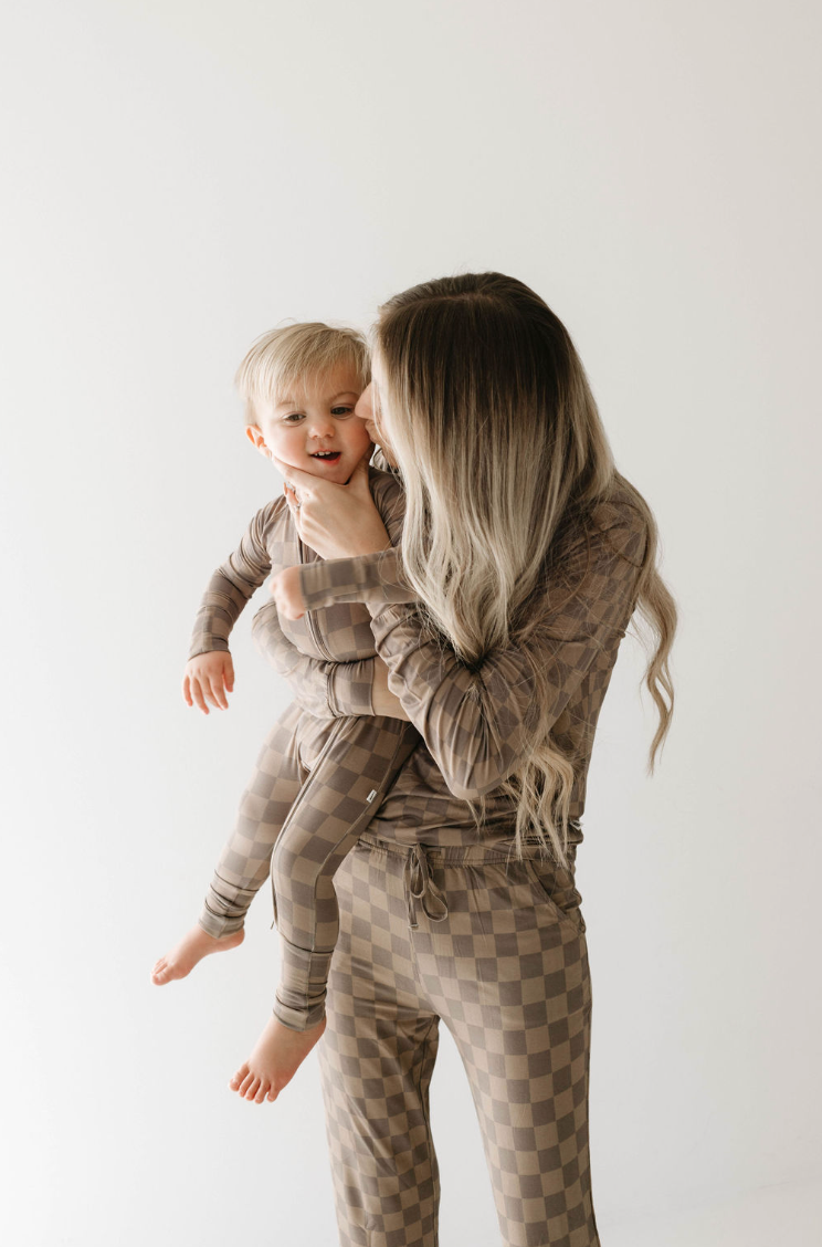 A woman with long, light hair cradles a young child in her arms. Both are dressed in matching outfits - Bamboo Zip Pajamas in Faded Brown Checkerboard by forever french baby. The setting is simple and clean, with a plain white background highlighting the hypo-allergenic bamboo clothing.