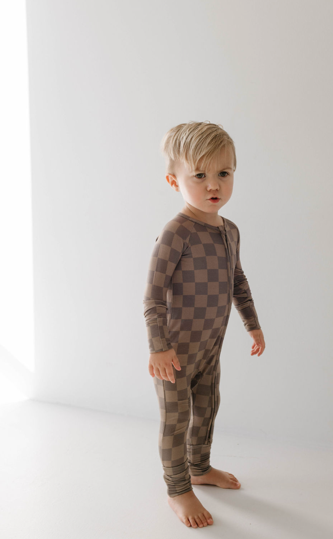A toddler with blonde hair stands indoors against a white background, wearing the "Bamboo Zip Pajamas | Faded Brown Checkerboard" by forever french baby. Made of breathable bamboo fabric, the one-piece outfit extends to cover their legs and arms. The child has a neutral expression on their face.