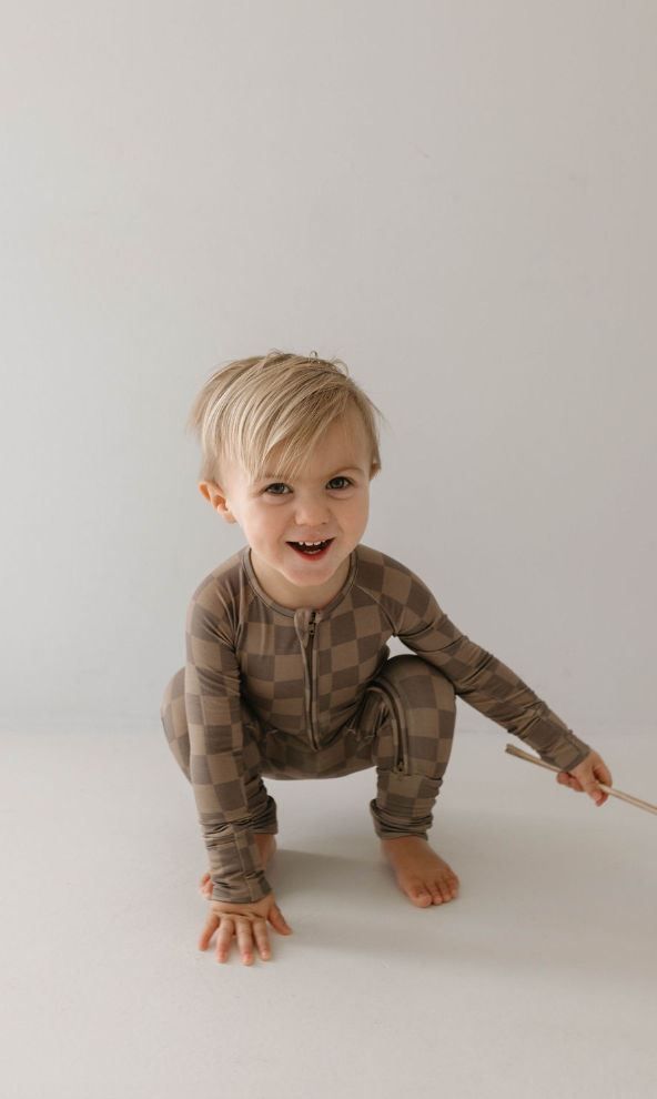 A young child with short blonde hair is smiling and kneeling on the floor. The child is wearing breathable and hypo-allergenic Bamboo Zip Pajamas in a Faded Brown Checkerboard pattern from forever french baby, and holding a stick. The background is plain and light-colored.