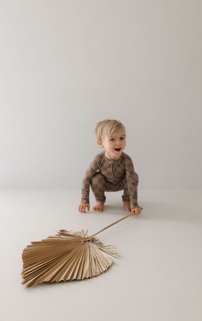 A young child with blond hair, dressed in the Forever French Baby Bamboo Zip Pajamas in Faded Brown Checkerboard, crouches on the floor with a large dried palm leaf in hand. The background is a simple, light-colored wall. The child is smiling and appears to be playing.