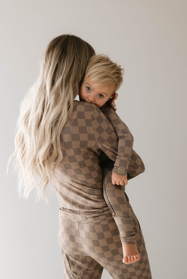 A person with long, wavy blonde hair is holding a small child. Both are wearing matching Bamboo Two Piece Pajamas in Faded Brown Checkerboard from forever french baby. The child, who has light blonde hair, is resting their head on the person's shoulder and facing the camera with a calm expression.