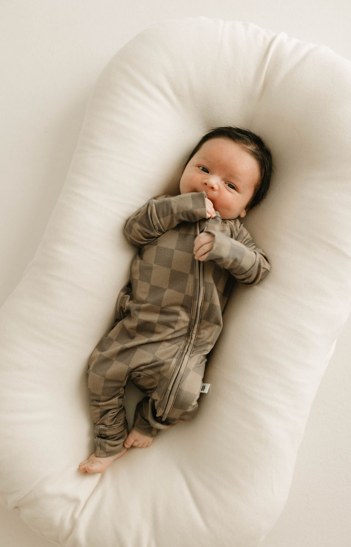 A baby with dark hair lies on a white cushioned surface, wearing the Bamboo Zip Pajamas in Faded Brown Checkerboard from forever french baby. The hypo-allergenic bamboo onesie feels comfortable as the baby rests peacefully with one hand near their mouth.