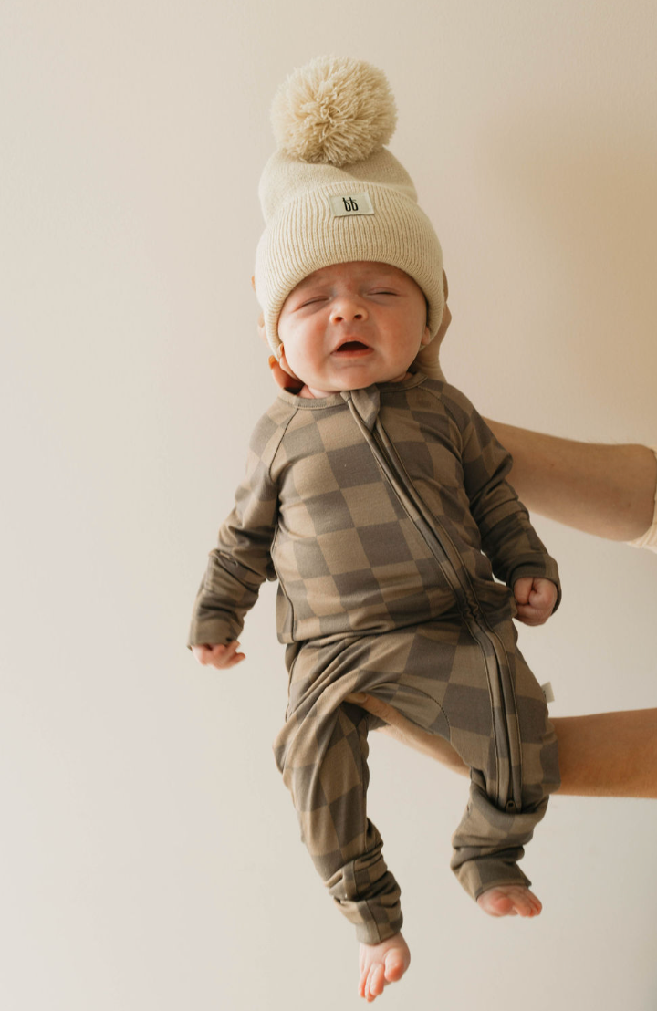 A baby dressed in Bamboo Zip Pajamas in Faded Brown Checkerboard by forever french baby, complemented by a cream knit hat with a large pom-pom on top, is being held up by an adult's hand against a plain beige background. The baby's expression seems to be squinting or grimacing.