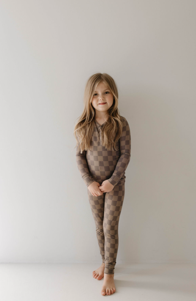 A young girl with long hair stands against a white wall. She is barefoot and dressed in hypoallergenic baby clothing from forever french baby, wearing the Bamboo Two Piece Pajama in Faded Brown Checkerboard. She smiles softly, with her hands clasped in front of her.